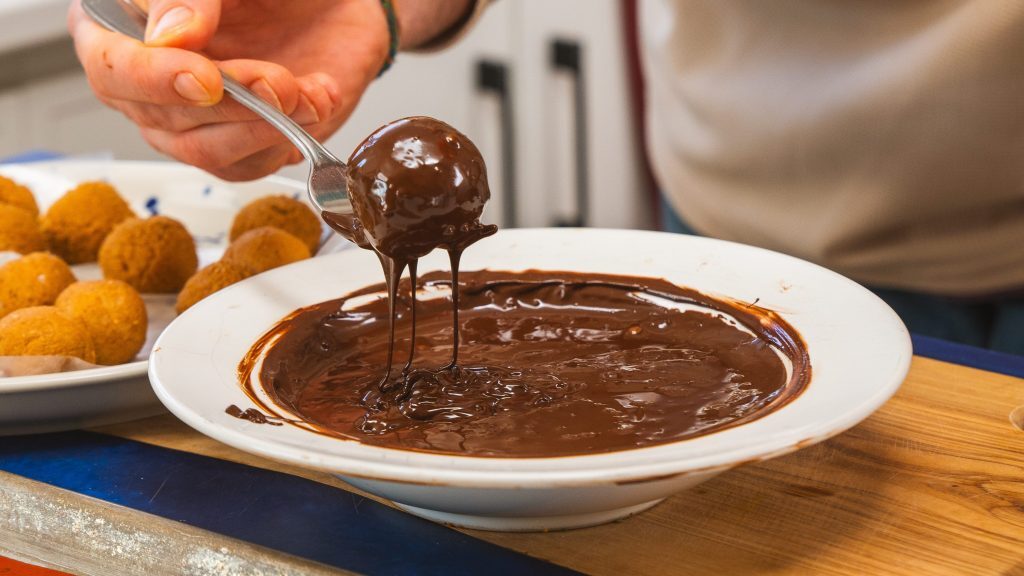 A white bowl filled with dar chocolate and a hand holding a spoon with a protein peanut butter ball that has just been immersed in the chocolate.