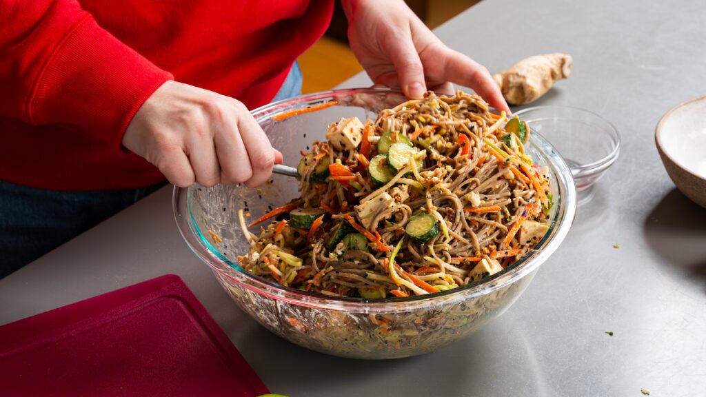 Soba noodle salad being tossed gently in a mixing bowl.