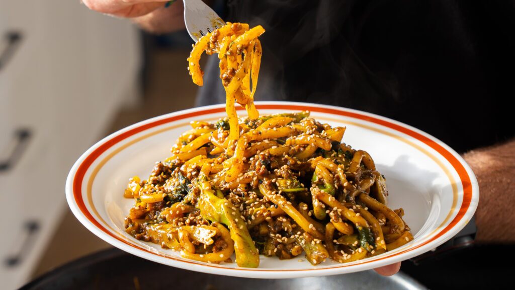 Close-up of a fork lifting a thick udon noodle from a serving bowl.