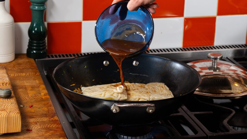 A dark brown sauce being added to a hot wok filled Fresh udon noodles.