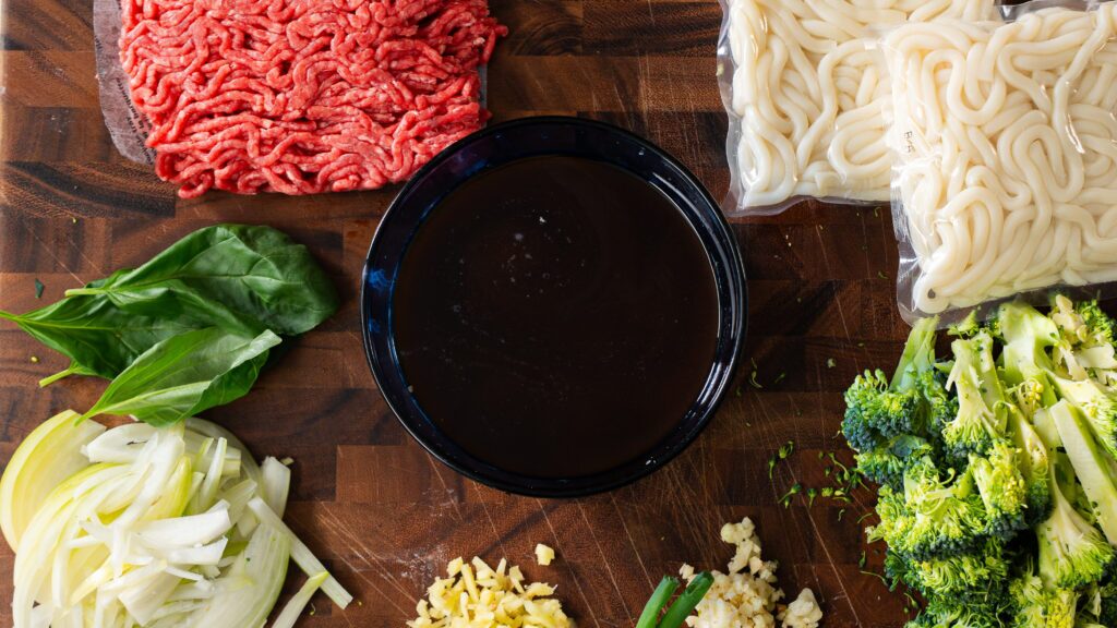 Chopped broccoli, basil leaves, and ground beef, green onions and other ingredients arranged on a cutting board.