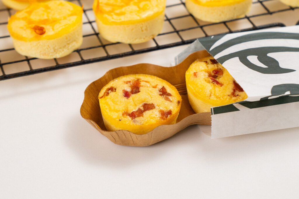 The image shows two bacon-filled Starbucks-style egg bites in a paper holder with more egg bites cooling on a rack in the background.