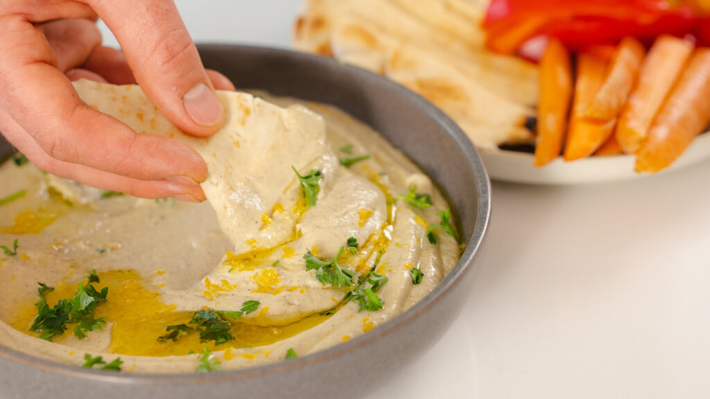 The image shows a hand dipping a piece of flatbread into a bowl of this creamy baba ganoush recipe, which is garnished with olive oil and chopped parsley. In the background, there's a plate with sliced vegetables and more flatbread.