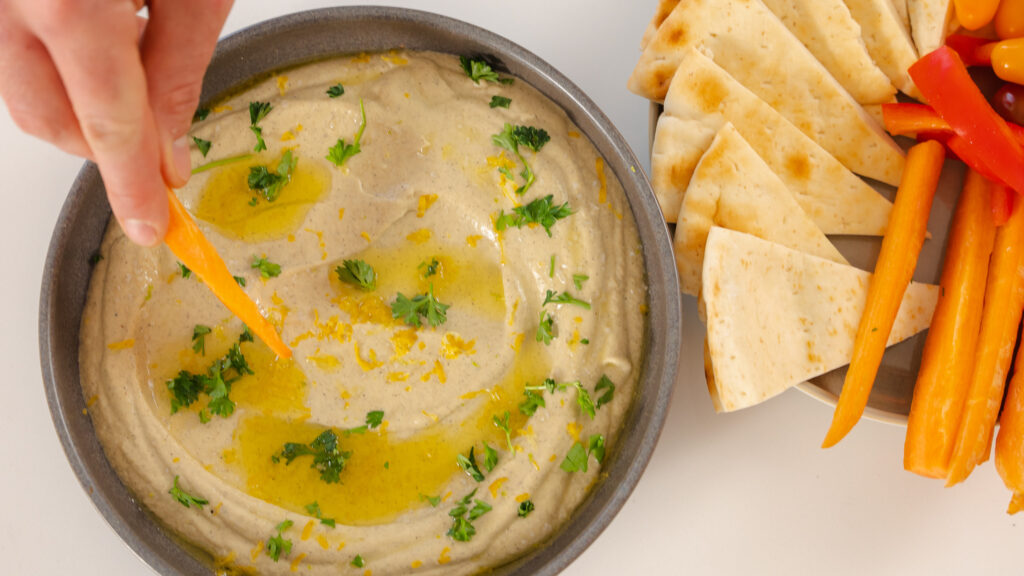 The image shows a hand dipping a piece of flatbread into a bowl of this creamy baba ganoush recipe, which is garnished with olive oil and chopped parsley. In the background, there's a plate with sliced vegetables and more flatbread.