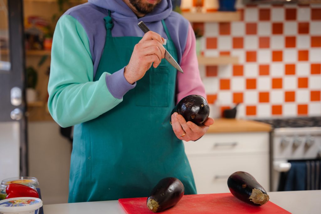 The image shows a person wearing a colorful hoodie and a teal apron, preparing to cook by making small incisions in an eggplant with a knife. The scene takes place in a kitchen with a red and white checkered backsplash. Two other eggplants rest on a red cutting board on the counter in front of them, suggesting the beginning of a cooking process, likely roasting the eggplants. 