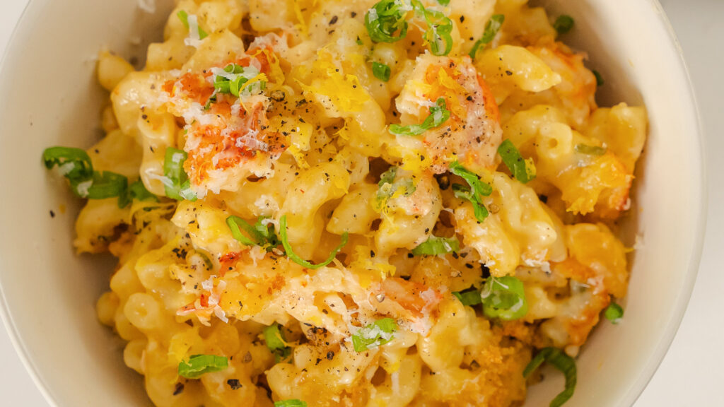 A close up of lobster mac and cheese. The beautiful red meat of the lobster is peeking though layers of yellow noodles with pops of green from freshly sliced green onion.