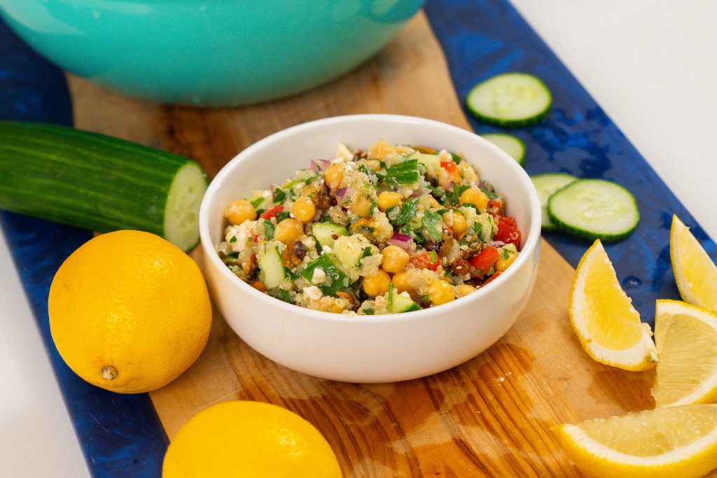 A white bowl filled with the Jennifer Aniston salad sits on a wooden board. The salad includes quinoa, chickpeas, cucumbers, red peppers, feta, and parsley, with slices of lemon and cucumber placed alongside for garnish.