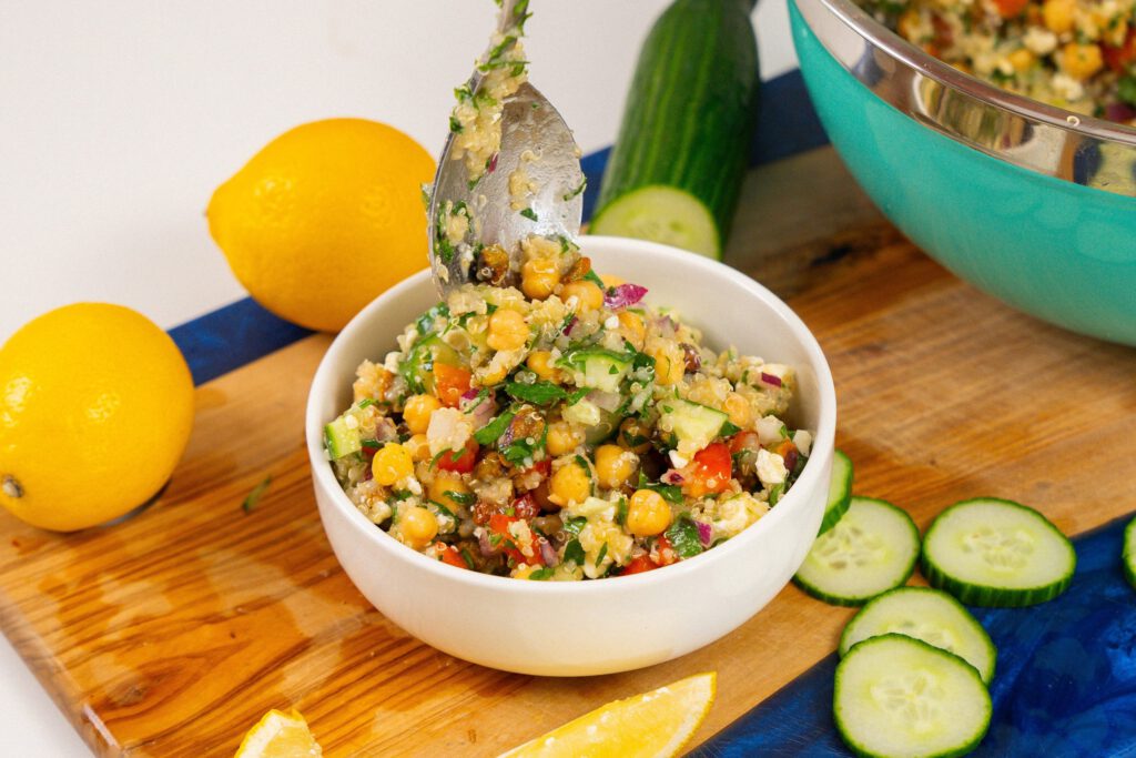 A spoon is lifting a portion of the Jennifer Aniston salad from a white bowl, showing a vibrant mix of chickpeas, quinoa, cucumber, red peppers, and feta. The bowl is placed on a wooden board, surrounded by lemon slices, whole lemons, cucumber slices, and a whole cucumber.