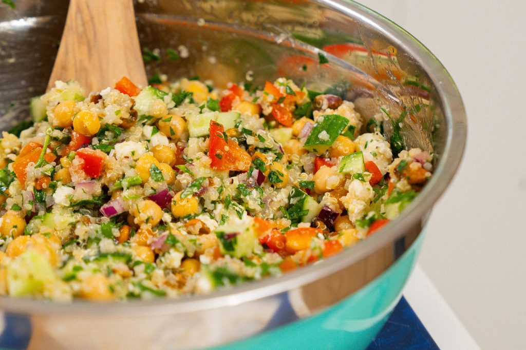 A close-up shot of the Jennifer Aniston salad in a large metal mixing bowl with a wooden spoon. The salad is a colorful mix of chickpeas, quinoa, cucumbers, red peppers, feta, and herbs, all tossed together for a fresh, vibrant appearance.