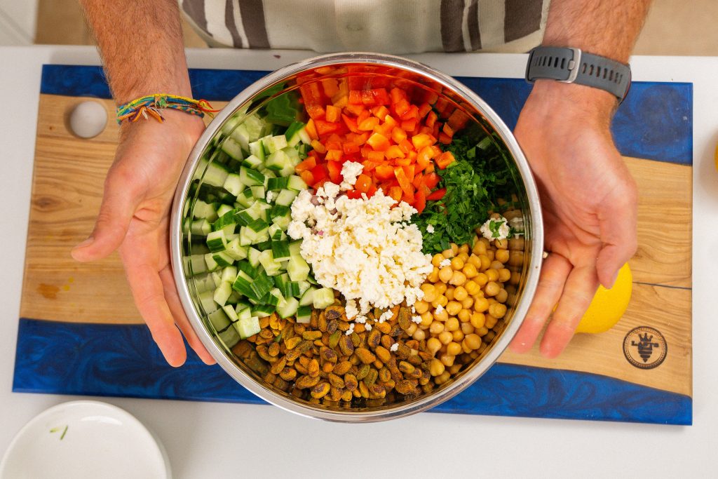 A pair of hands holds a large mixing bowl filled with the separate ingredients for the salad. The bowl displays neat sections of diced cucumber, red peppers, chickpeas, crumbled feta, chopped parsley, and pistachios, ready to be mixed together.