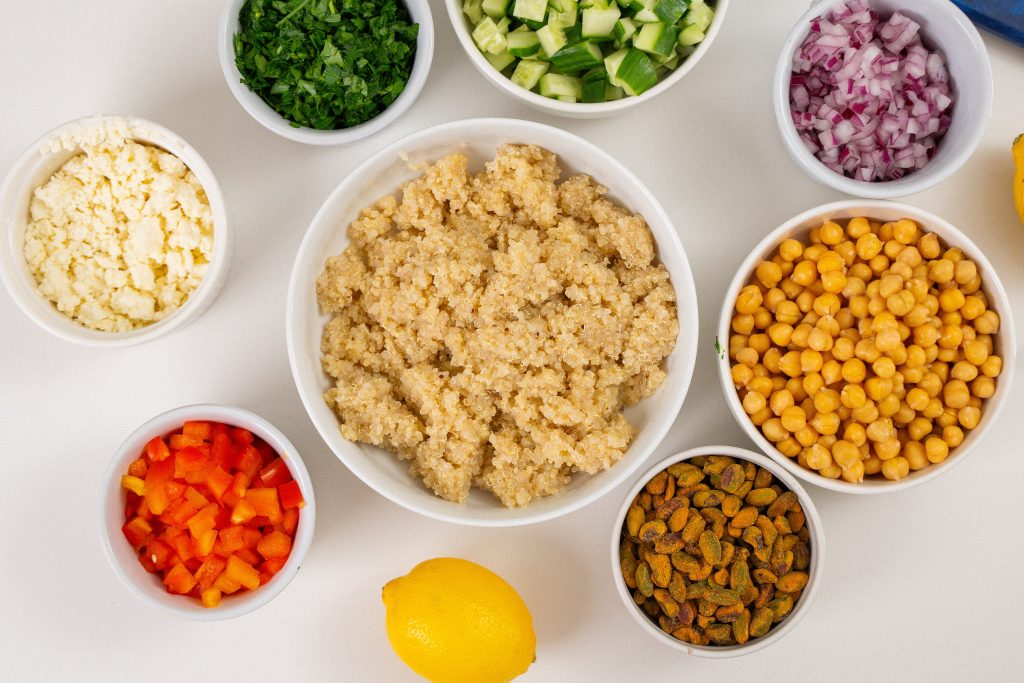 The photo shows a variety of small bowls filled with ingredients arranged on a white surface. In the center is a bowl of cooked quinoa, surrounded by bowls containing chopped red peppers, crumbled feta, chickpeas, pistachios, parsley, diced cucumber, and red onion, with a lemon placed in front.