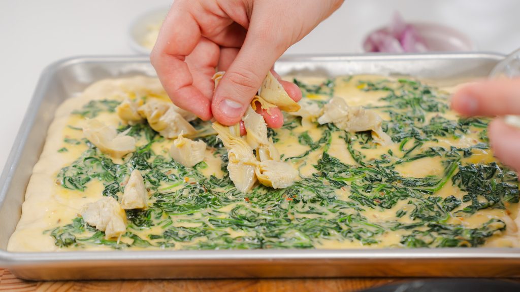 In the process of making this Spinach and Artichoke Veggie Pizza. A hand is gently laying sliced artichokes onto a unbaked pizza with a layer of creamy green spinach.  