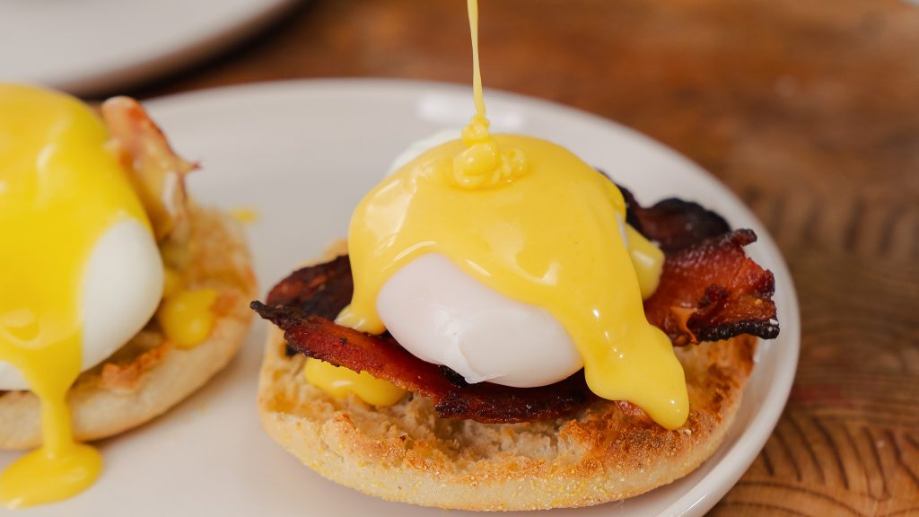 Hollandaise sauce being poured over a poached egg on top of an english muffin. 