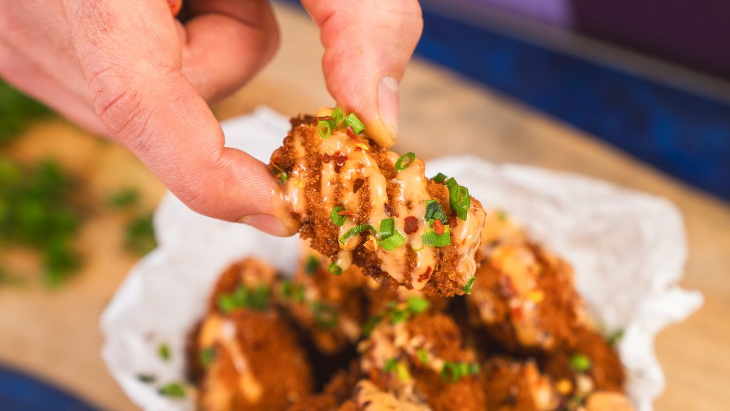 A close up shot of Andy holding a little golden nugget of Bang Bang Chicken. It's covered in a orange sauce with green chives. 