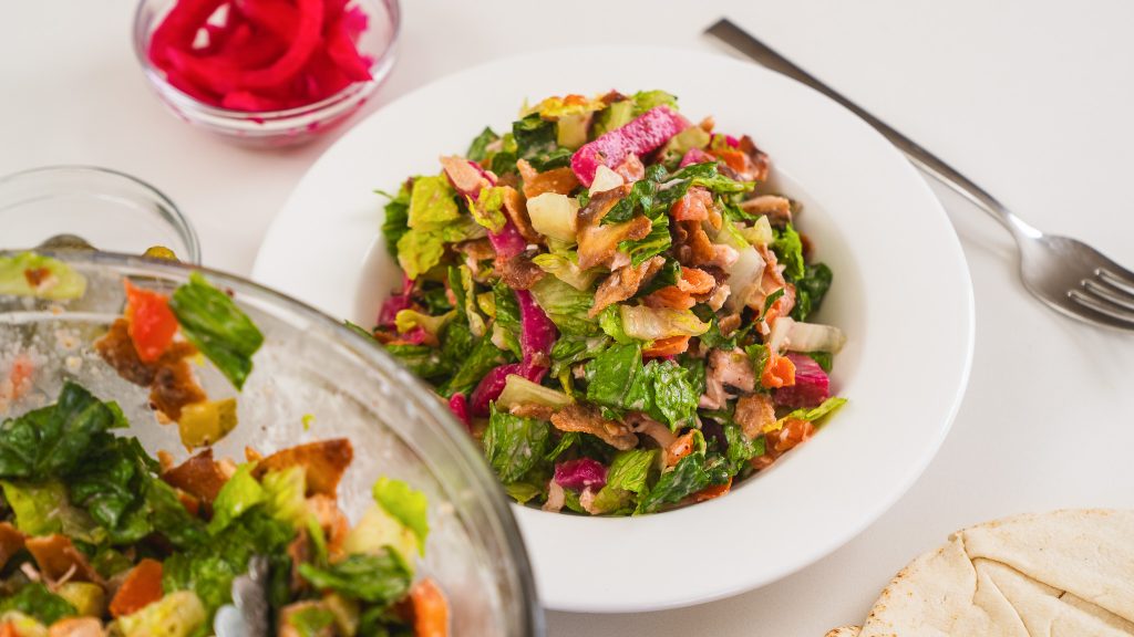 A close up shot of Chicken Shawarma Salad in a white bowl surrounded by it's ingredients.