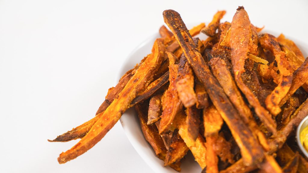 A close up shot of Sweet Potato Fries in a white bowl on a white counter. They are bright orange with golden brown flecks. 