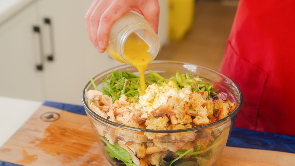 A close up shot of Garlic Lemon Dijon Vinaigrette beinfgpoured from a mason jar into to a salad of greens, chicken and toasted croutons.