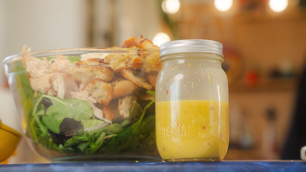 A close up shot of Garlic Lemon Dijon Vinaigrette in a half full mason jar next to a salad of greens, chicken and toasted croutons.