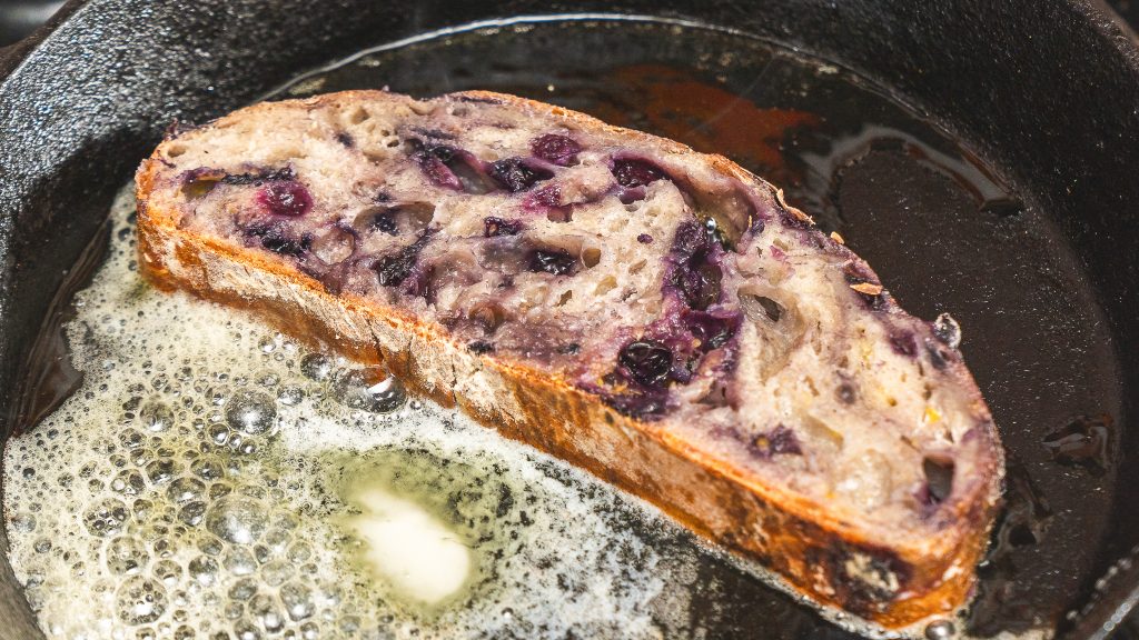 The image shows a slice of no knead bread frying in butter in a cast-iron skillet, creating a golden, crisp texture. The blueberries are visible within the bread as it cooks.