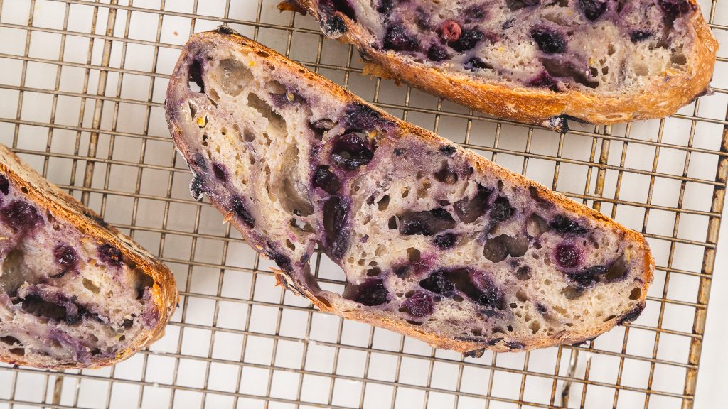 The image shows slices of lemon blueberry bread on a cooling rack, revealing a rustic texture with swirls of blueberries throughout the bread. The crust is golden and the interior is studded with vibrant blueberries.