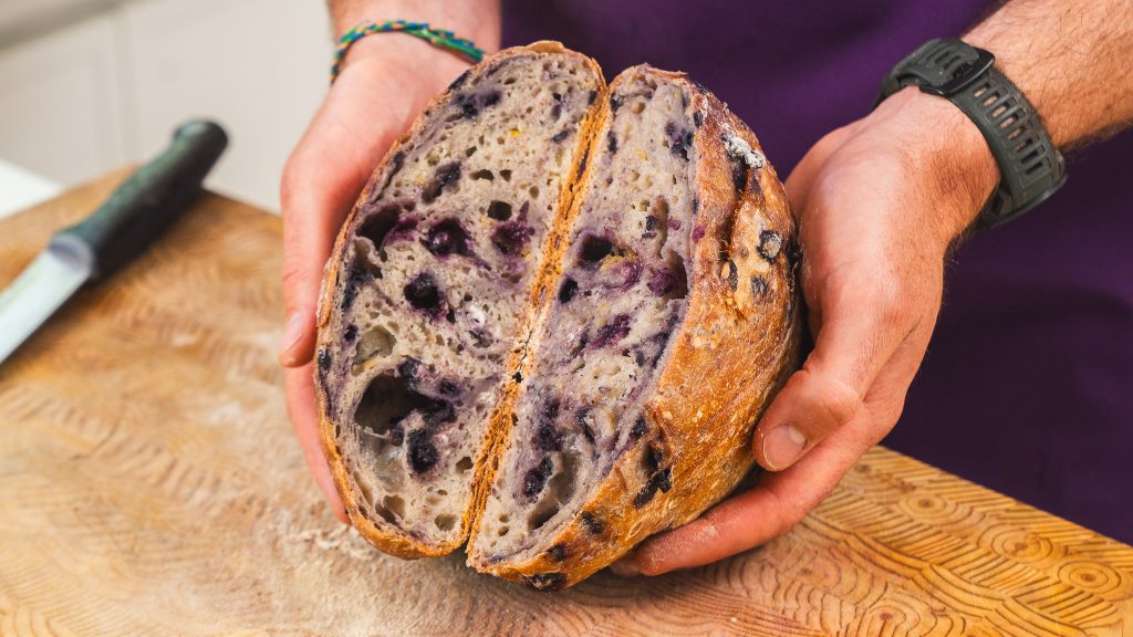 The image shows a person holding a loaf of lemon blueberry bread, sliced in half, revealing a soft and airy crumb filled with blueberries. The crust has a golden-brown, rustic look.