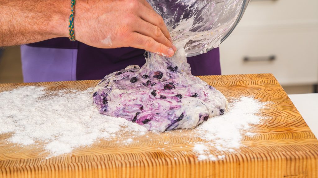 The image shows a person transferring dough from a bowl onto a floured surface. The dough is soft and sticky