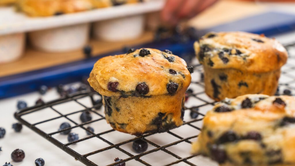 close up image of blueberry muffin sitting on a cooling rack. Loose blueberries are underneath the cooling rack