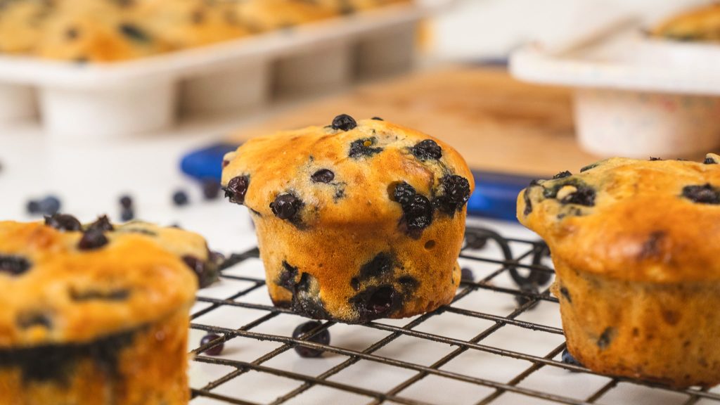 close up image of blueberry muffin sitting on a cooling rack. Loose blueberries are underneath the cooling rack