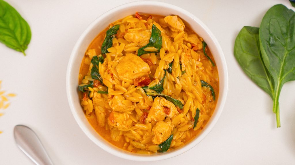 A top-down view of a bowl of Marry Me Chicken Orzo, showcasing the creamy orzo, chicken chunks, spinach, and sun-dried tomatoes, with fresh basil leaves nearby