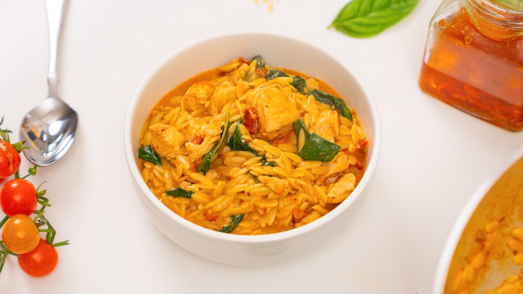 A close-up of a white bowl filled with creamy Marry Me Chicken Orzo, with vibrant spinach leaves and tomatoes placed next to the bowl, creating a fresh and appetizing presentation