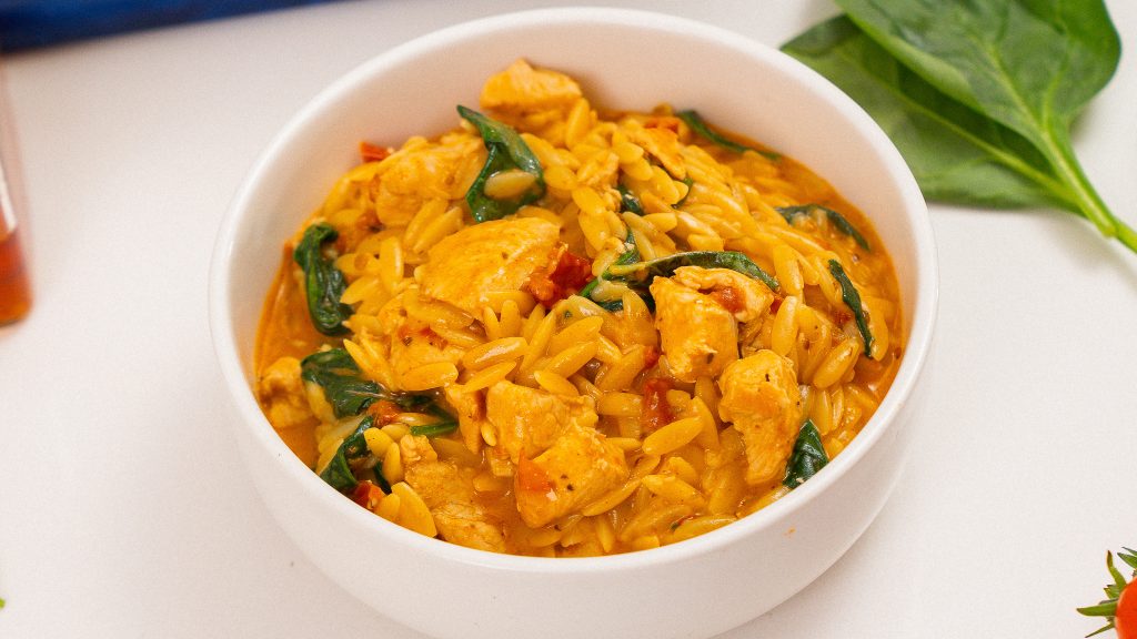 A close-up of a white bowl filled with creamy Marry Me Chicken Orzo, with vibrant spinach leaves and tomatoes placed next to the bowl, creating a fresh and appetizing presentation