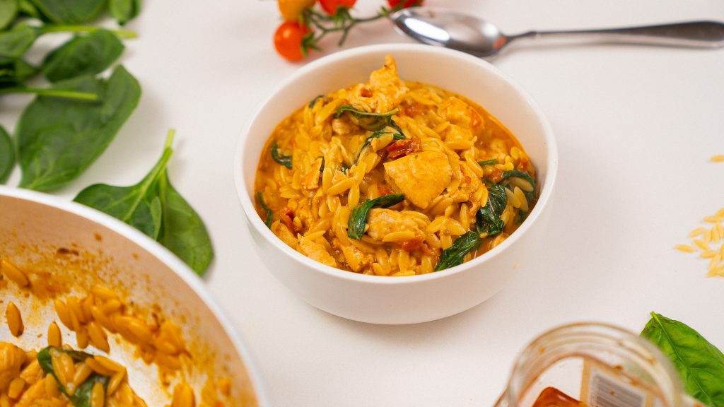 A white bowl of creamy Marry Me Chicken Orzo, loaded with tender chicken, sun-dried tomatoes, and spinach, with fresh spinach leaves and cherry tomatoes scattered in the background.