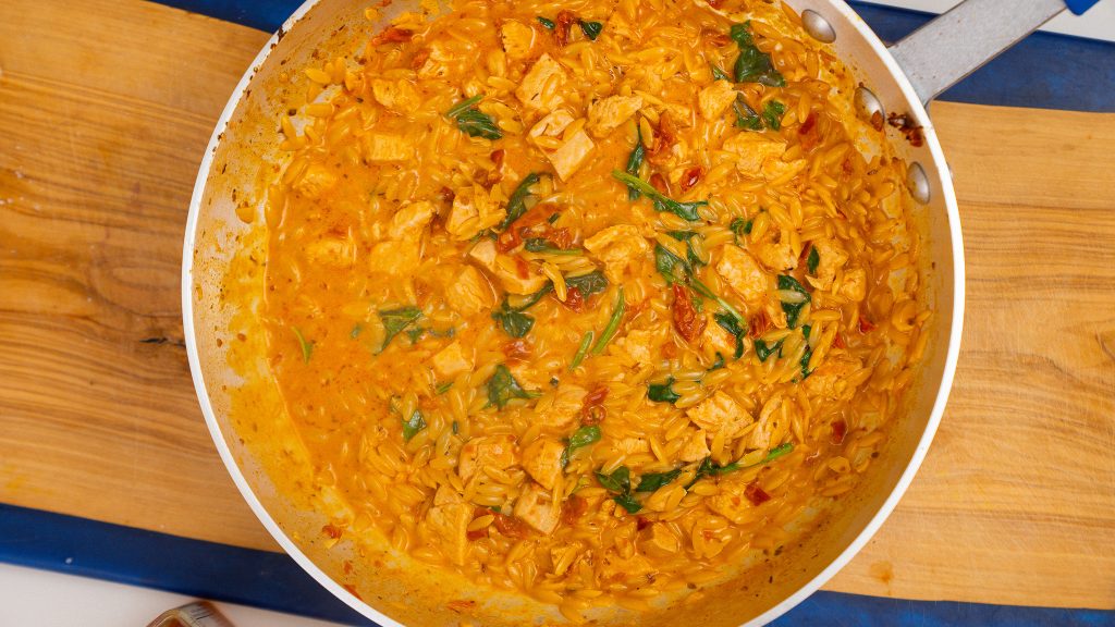 A pan filled with creamy Marry Me Chicken Orzo, garnished with fresh spinach and sun-dried tomatoes, with a wooden cutting board in the background.