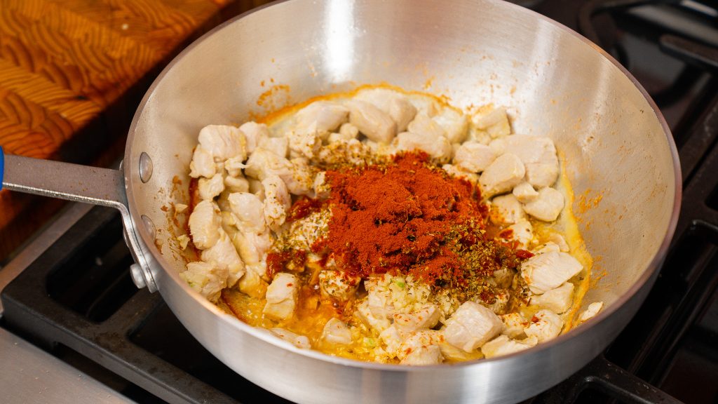 Chicken pieces being sautéed with garlic, paprika, and oregano in a stainless steel pan on a stove, starting the base for the Marry Me Chicken Orzo recipe