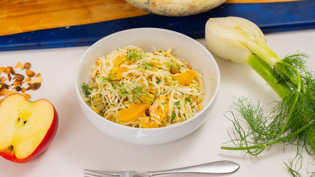 45 degree angle of bowl of orange tahini slaw. Apple and fennel bulb on each side of bowl