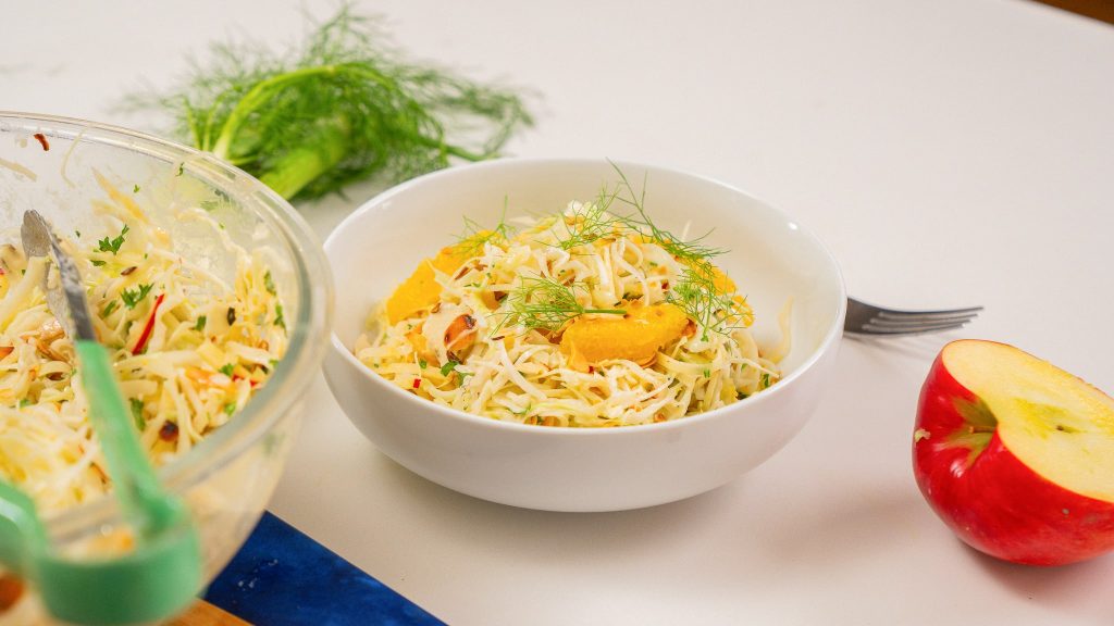 side angle view of orange fennel slaw. This cabbage salad is topped with oranges, and fennel. Glass bowl of salad off to the left, fennel in back ground