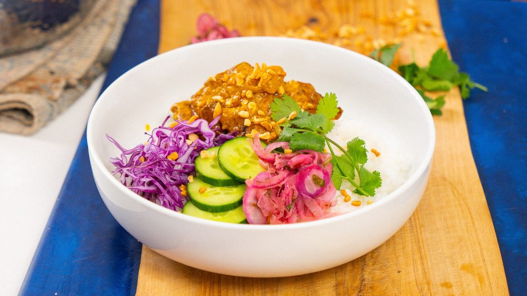 Satay chicken rice bowl served with pickled red onions, sliced cucumbers, shredded purple cabbage, and fresh cilantro, garnished with chopped peanuts