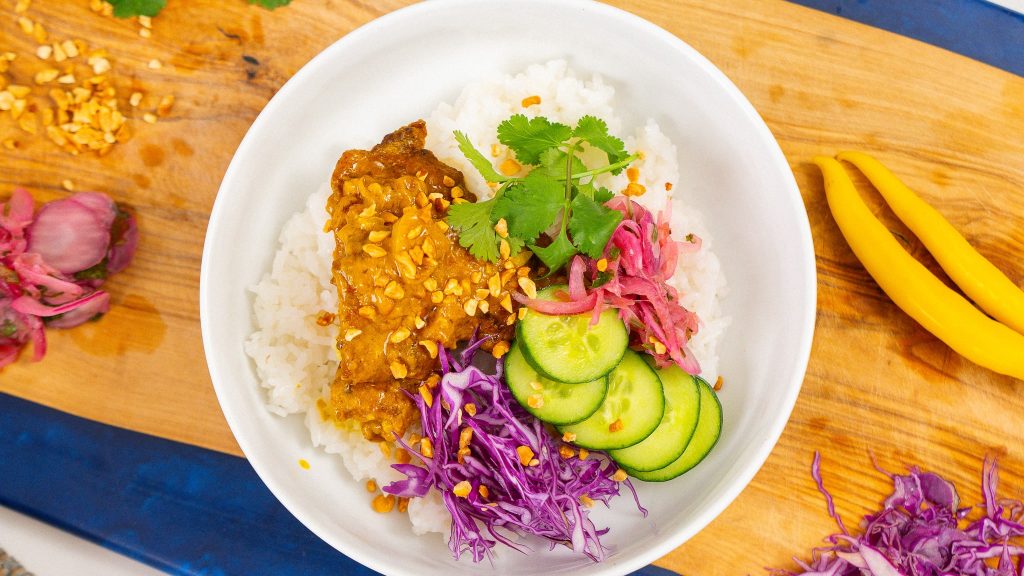 Overhead view of a satay chicken rice bowl topped with peanut sauce, pickled red onions, sliced cucumbers, shredded purple cabbage, and fresh cilantro, garnished with chopped peanuts