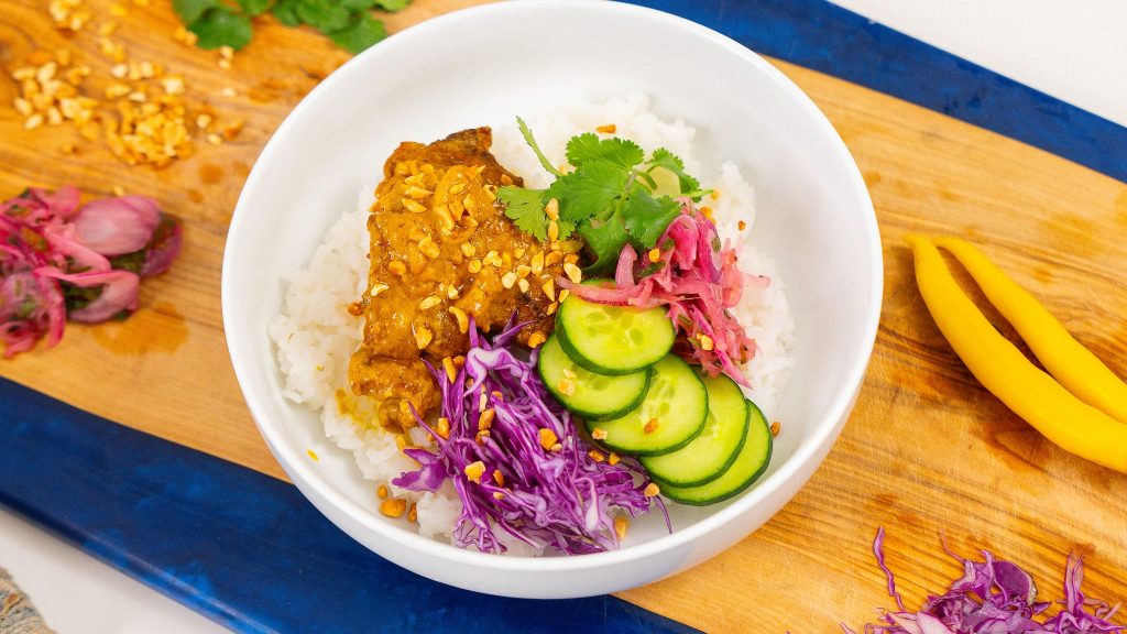 Satay chicken rice bowl topped with peanut sauce, sliced cucumbers, pickled red onions, shredded purple cabbage, and chopped peanuts, served on a bed of white rice with fresh cilantro garnish