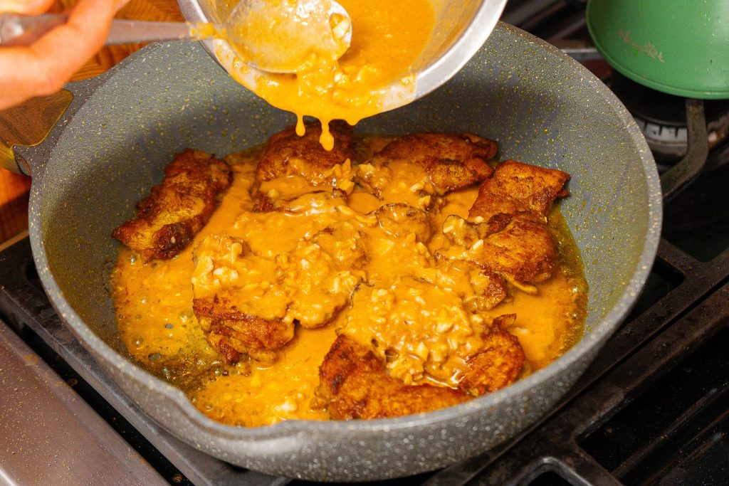 Pouring peanut sauce over seared chicken thighs cooking in a pan, creating a rich and flavorful satay chicken dish