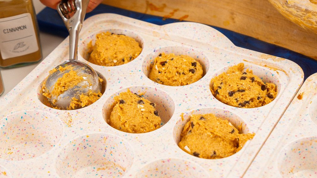 Batter being portioned into a speckled muffin tray using a metal scoop, with cinnamon jars in the background