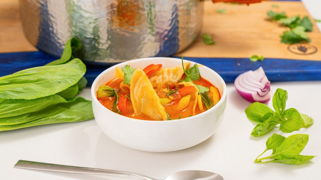 A close-up of a bowl of red curry dumpling soup, featuring a dumpling floating in the bright red broth, with fresh cilantro and basil garnishing the top.