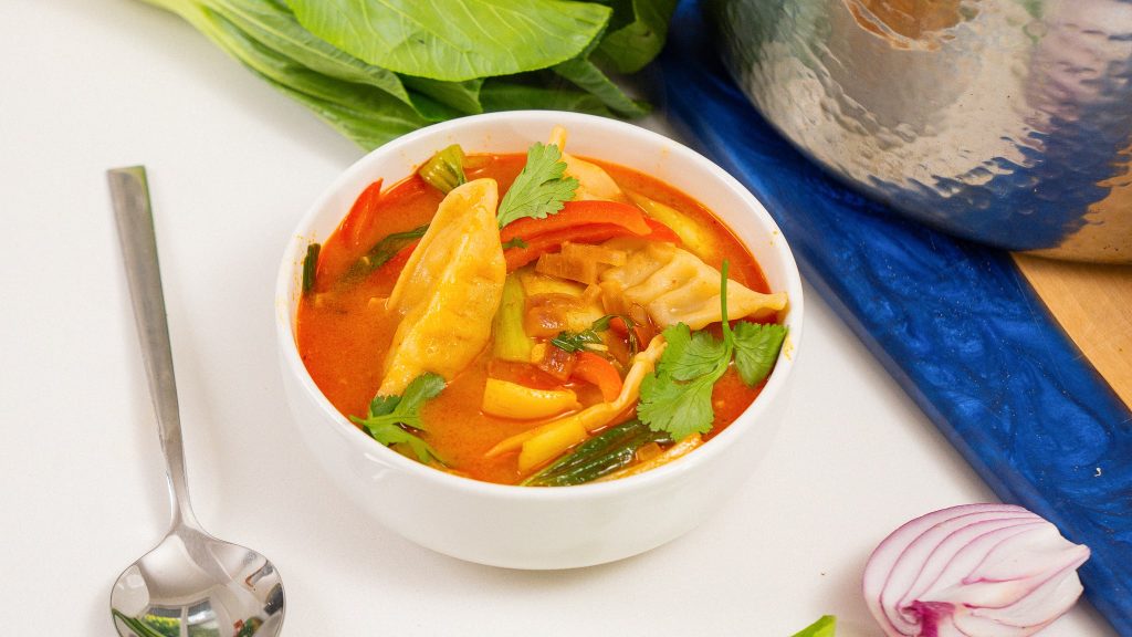 A bowl of red curry dumpling soup with baby bok choy, red bell peppers, and fresh herbs, next to a spoon and a cutting board.