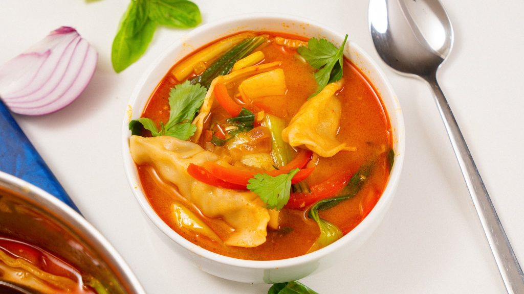 A bowl of red curry dumpling soup with baby bok choy, red bell peppers, and fresh herbs, next to a spoon and a cutting board.