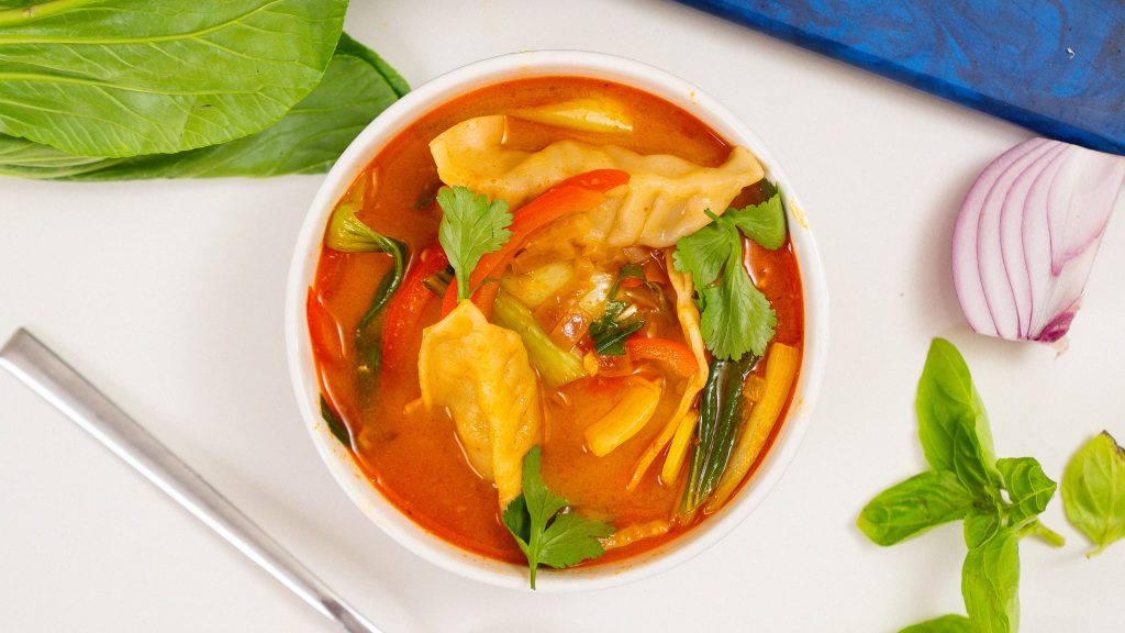 A top-down view of a bowl filled with red curry dumpling soup, garnished with fresh basil and cilantro, surrounded by fresh vegetables and herbs.