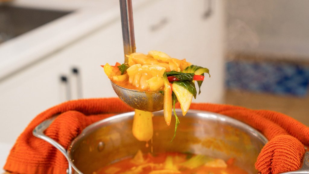 A ladle filled with dumplings and vegetables being lifted from a pot of red curry soup, with steam rising.