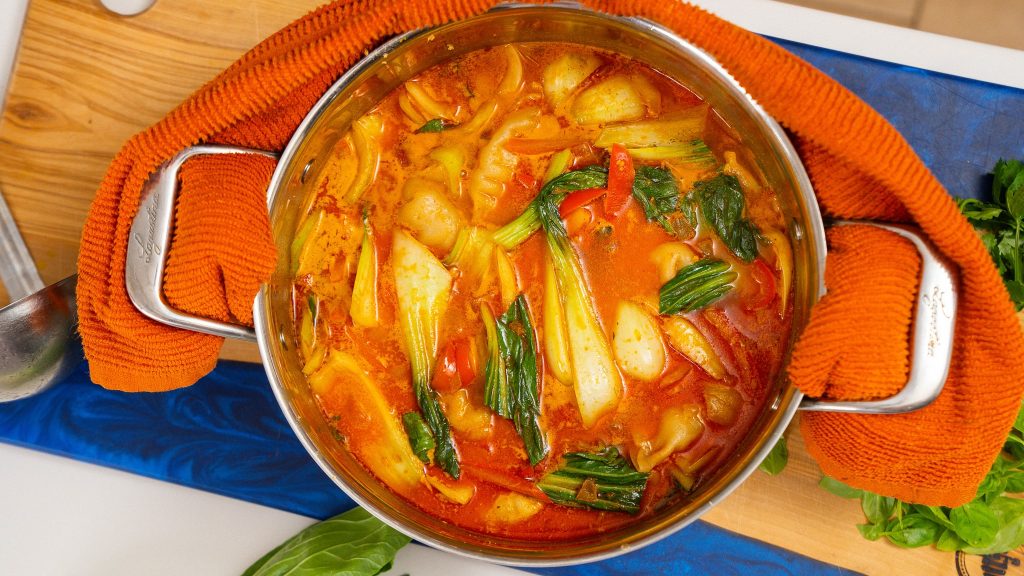 A close-up of a simmering pot of red curry broth with bok choy, bell peppers, and dumplings visible, surrounded by fresh herbs.