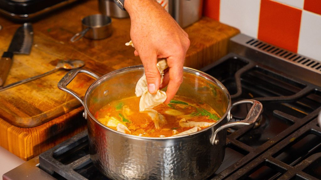 A hand dropping frozen dumplings into a simmering pot of red curry broth on the stove.