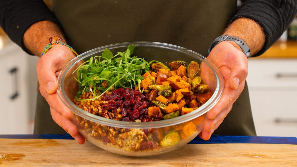 hands holding a clear bowl with sweet potatos, cranberries, arugula