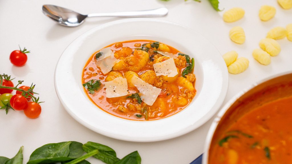 Chicken sausage gnocchi soup served in a white bowl, with spinach and cherry tomatoes artfully arranged around the dish for a fresh, vibrant look.